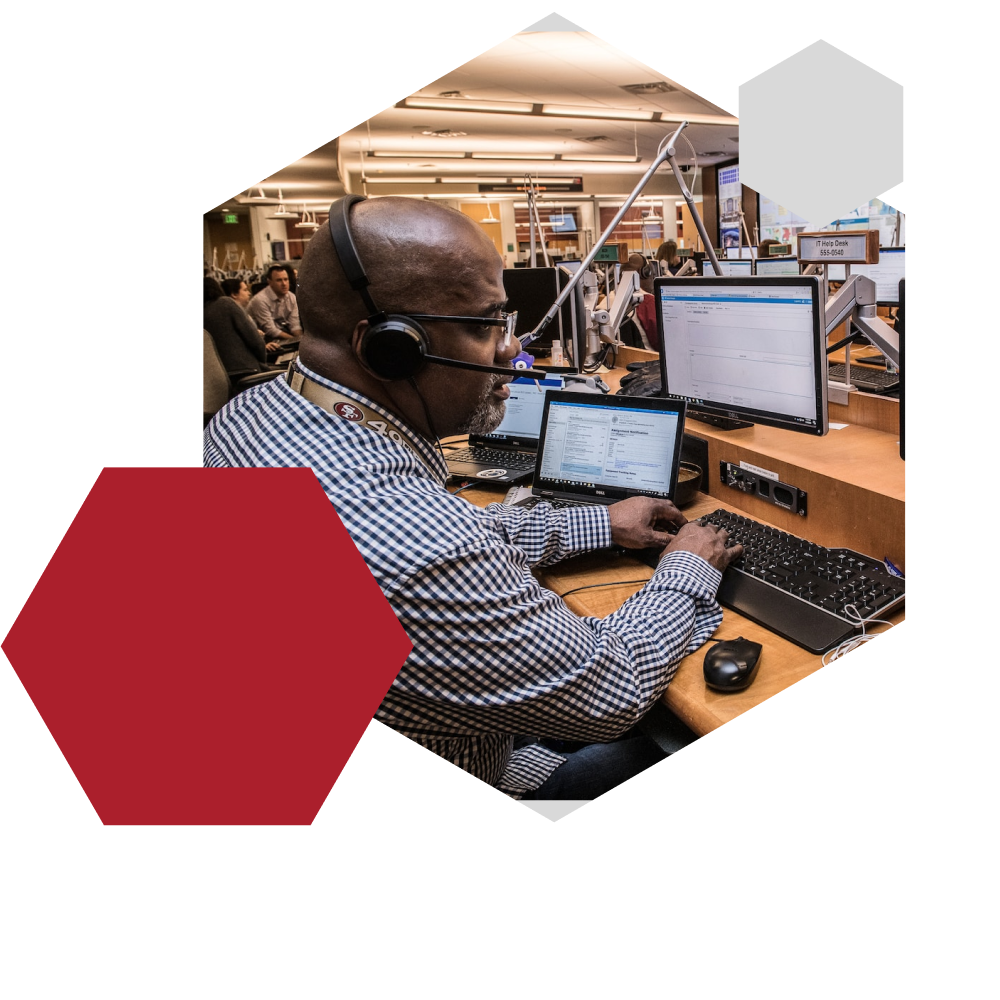 A communications specialist wearing a headset works at a desk with multiple monitors and a laptop in a busy office environment. The workspace includes advanced tech tools and other team members in the background, reflecting Governor Communications’ extensive network of partners across Jamaica and internationally. Framed in a hexagonal design with red and white overlays, the image highlights real-time collaboration, customer engagement, and global connectivity.