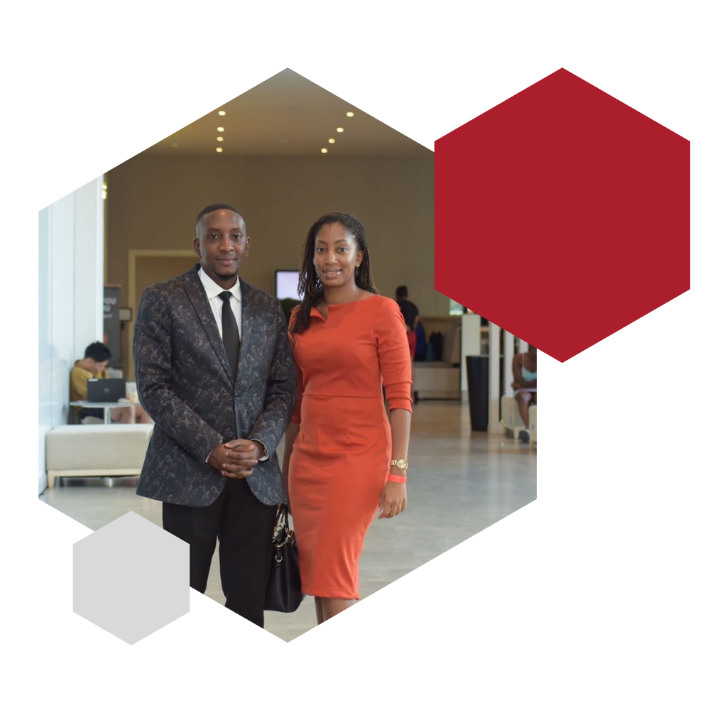 Joseph Cunningham, CEO of Governor Communications, stands in a dark patterned suit beside a woman in a fitted red dress at a formal indoor event. The venue features high ceilings, recessed lighting, and white furnishings, with other guests visible in the background. Framed in a hexagonal design with red and light gray overlays, the image reflects Governor Communications’ commitment to professional representation, public relations excellence, and brand leadership in Jamaica.