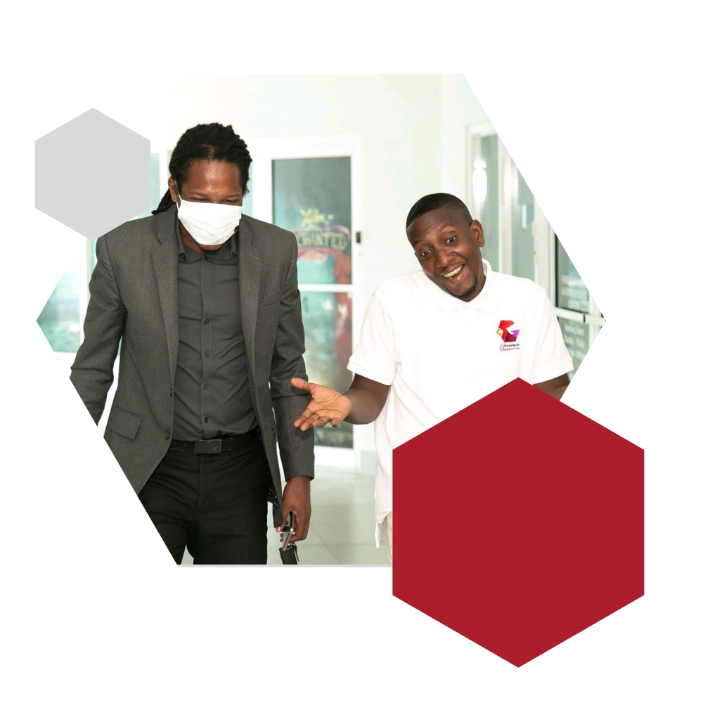 Joseph Cunningham, CEO of Governor Communications, gestures while smiling during a conversation with Jamaican politician Damion Crawford, who wears a grey blazer, black shirt, black pants, and a white face mask while holding a laptop. The two walk through a bright, modern office interior framed by hexagonal graphics in red and grey. The image reflects Governor Communications’ commitment to strategic leadership, public engagement, and collaborative innovation.